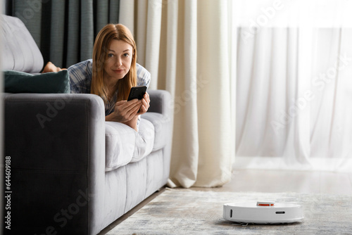 The girl lies on the sofa in the living room and controls a smart robot vacuum cleaner using a smartphone.