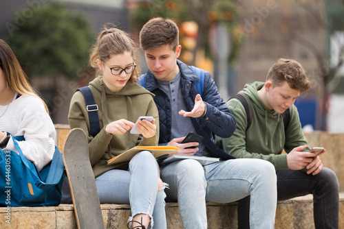 Group of boys and girls teenager addicted in their phones sitting on bench outdoors