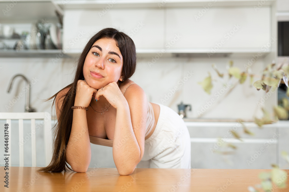 Positive young woman posing cheerfully leaning on table in the kitchen with white furniture