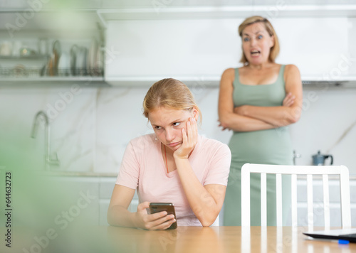 Upset teenage girl immersed in phone sitting at home table while disapproving mother standing in background, reflecting concern over daughter detachment from environment
