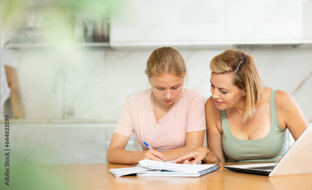 Fototapeta premium Mom prompts child, monitors implementation of lessons, homework on school subjects. Woman looks at laptop screen and at notebook of teenage daughter
