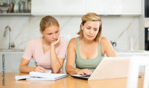Positive woman offering assistance and support to teenage daughter absorbed in writing notes in study book while sitting at table with laptop..