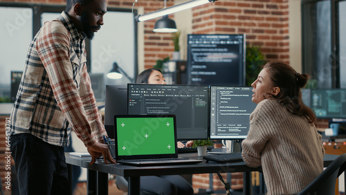 Programer working at desk with multiple computer screens and laptop with green screen chroma key mockup running code. Software developer compiling source algorithm interrupted by colleague.