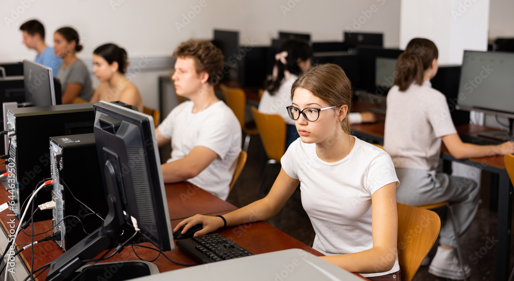 Teenage girl in glasses sitting at table and using computer during lesson.