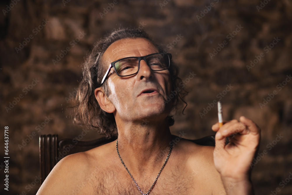 Fototapeta premium An older man, about 60 years old, of Serbian nationality with long hair and glasses, thoughtfully smokes a cigarette on a dark background. Artist, creative person. Happy face.