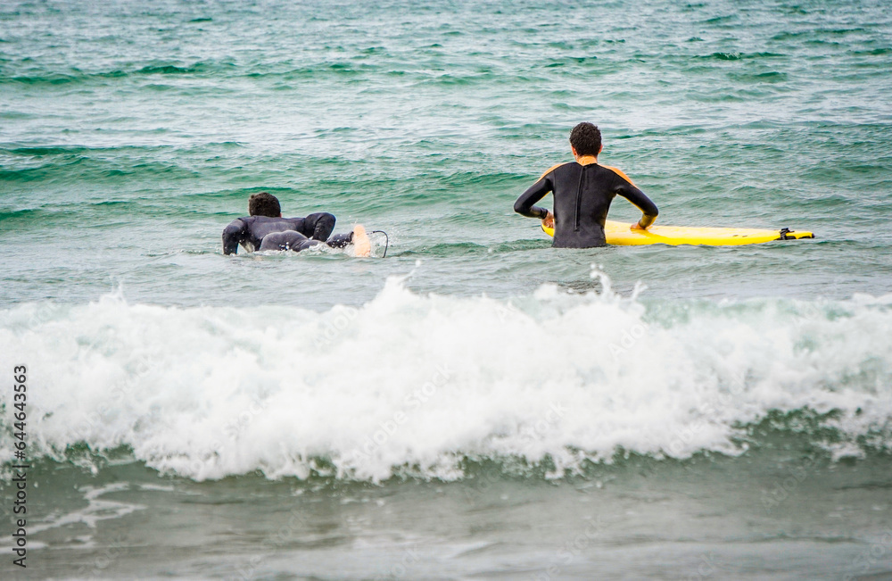 Two Surfers with Wetsuits seen from Behind Entering the Sea with their Boards with Wave Foam in Close Up