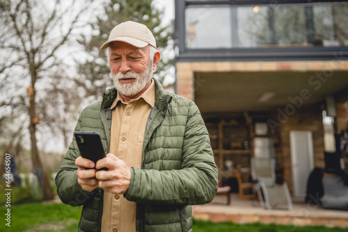 Obraz na plátne One senior man stand in front of tiny house in day use smart phone