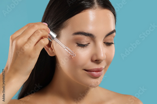 Beautiful young woman applying serum onto her face on light blue background, closeup