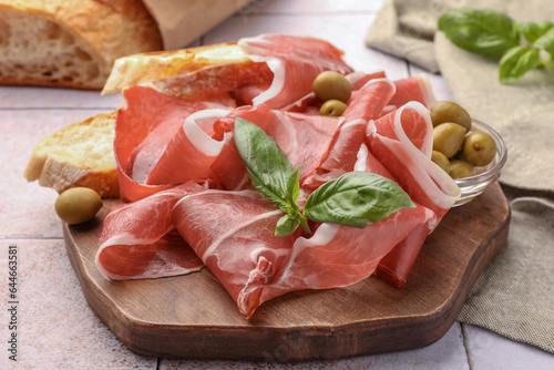 Slices of tasty cured ham, olives, bread and basil on tiled table, closeup