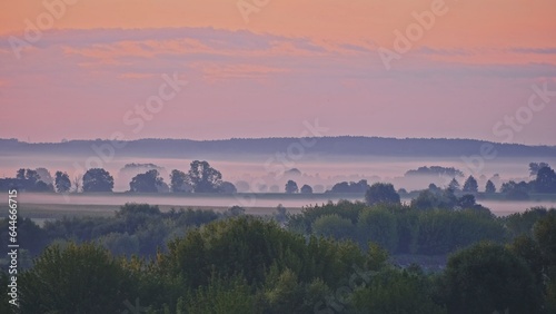 Beautiful Scenic Sunrise Morning Landscape with Fog Floating over River and Fields and Wind Turbines Rotating on Distatnt Hills on the Horizon 
