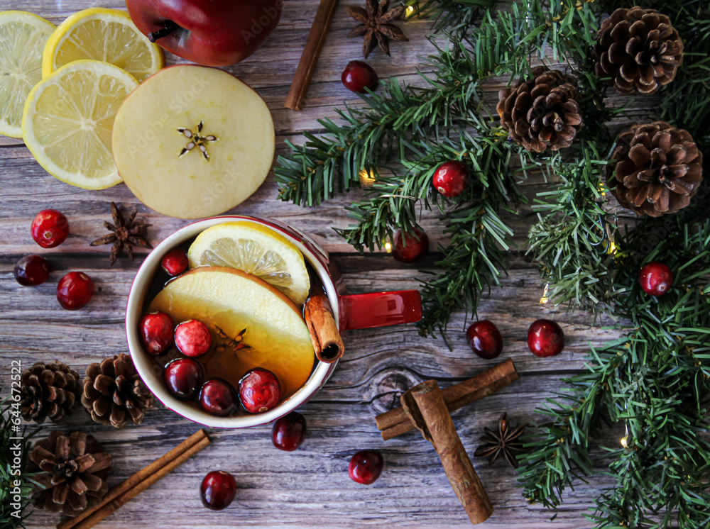 Christmas background flat lay with hot tea infused with fresh apple, lemon, cinnamon and cranberries on a textured wooden surface and green garland for Christmas spirit.