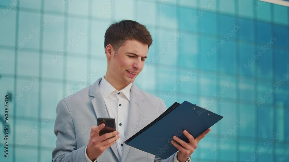 A young male engineer or architect in a formal suit during a coffee break stands near a building with blue windows and talks on the phone A young male engineer or architect in a formal suit during a coffee break stands near a building with blue windows and talks on the phone