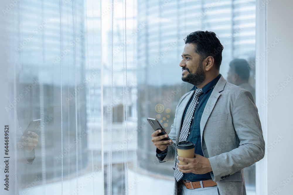 Indian businessman standing next to window using mobile phone wile coffee break Indian businessman standing next to window using mobile phone wile coffee break