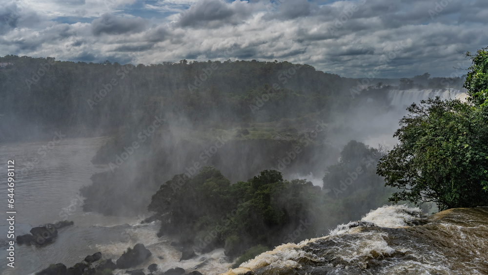 A stream of seething water collapses from a ledge. Spray and mist in the air. Lush tropical vegetation all around. Clouds in the sky. Argentina. Iguazu Falls.