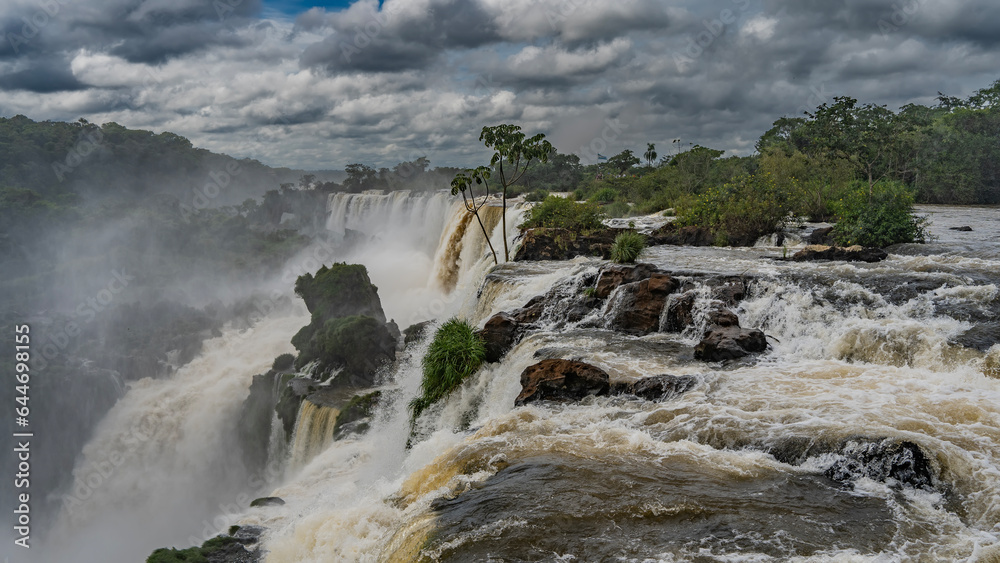 A stream of swirling water rushes along the rocky riverbed and collapses into the abyss. Green vegetation on the stones. Spray, fog all around. Clouds in the sky. Iguazu Falls. Argentina.