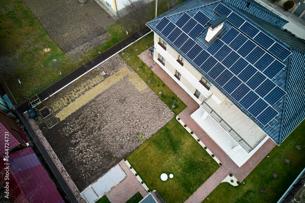 Aerial view of residential autonomous house with photovoltaic solar panel modules on rooftop. Home with solar electric system, lawn and pavement outside. Renewable energy and housing concept.