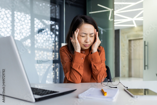 Overtired and overworked asian woman at workplace inside office, worker has severe headache and migraine, business woman holding hand on head.