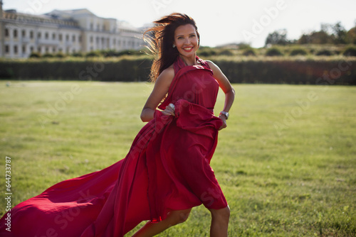 Brunette woman in long, flowing red dress running through park in nature, looking in camera, and smiling. Happy female model with long hair outdoors