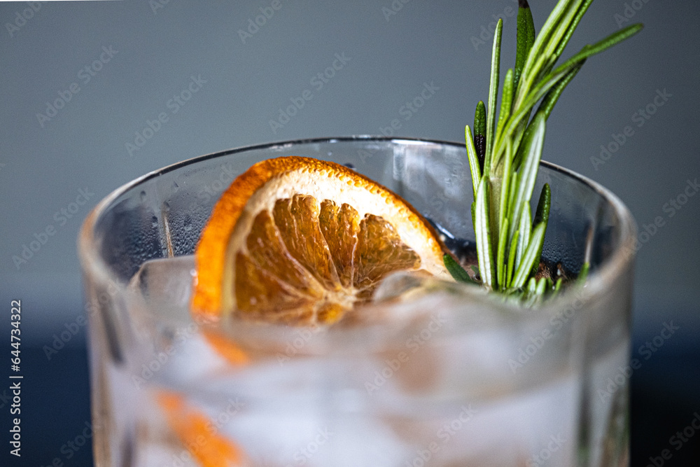 Glass of gin and tonic cocktails with ice, sprig of rosemary and sliced dry orange on a blue background. Copy space - obrazy, fototapety, plakaty 