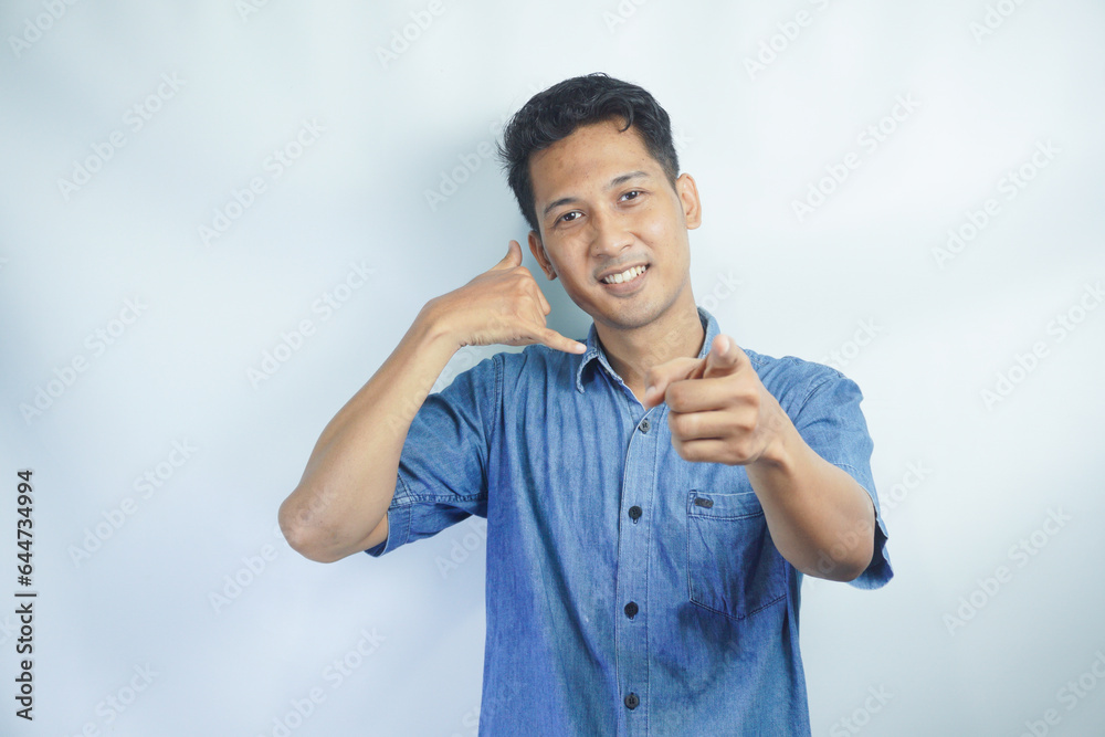 Fototapeta premium Young Asian man wearing blue shirt standing over isolated white background smiling doing talking on the telephone gesture and pointing to you. Call me.