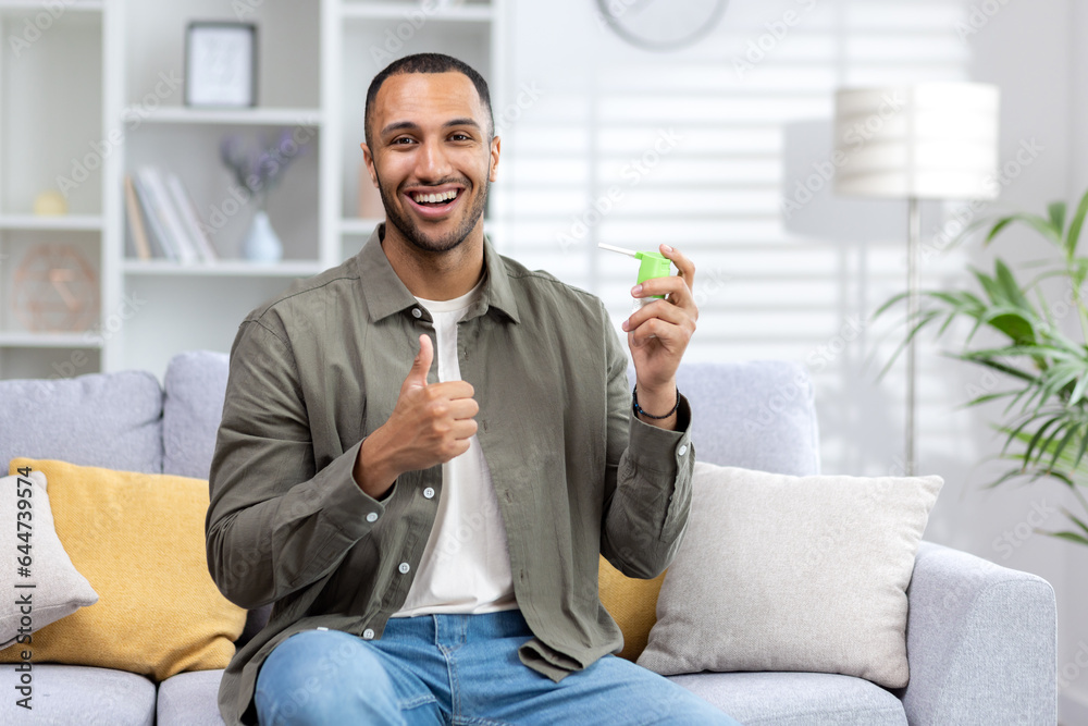 Portrait of a young African American man sitting on the couch at home and holding a throat spray in his hands and showing a super gesture with his finger - obrazy, fototapety, plakaty 