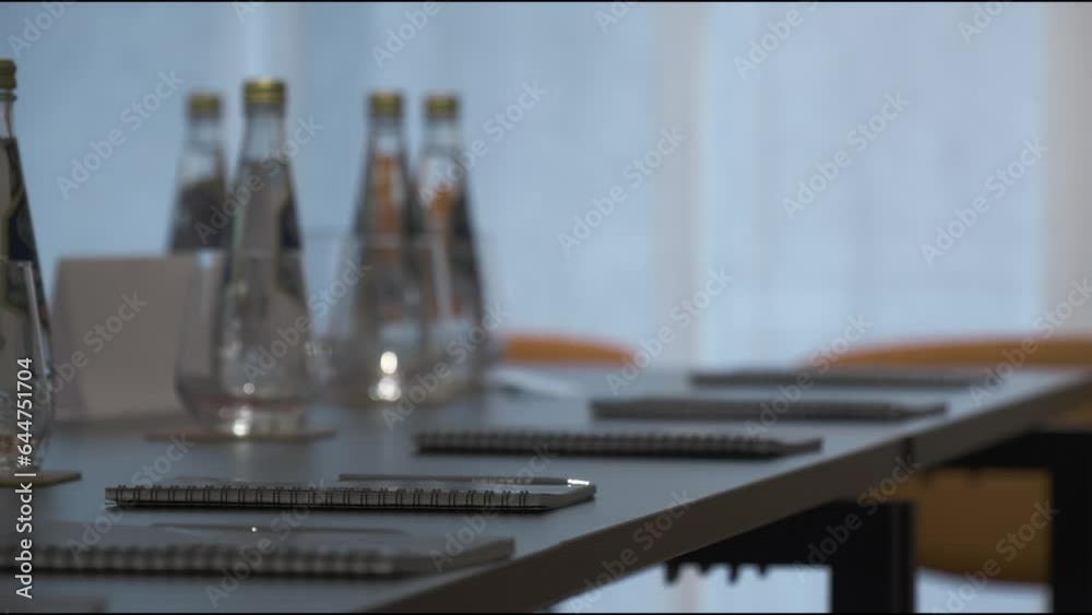 Assistant puts pens near notebooks on long conference table in office closeup. Woman prepares supplies for business forum in hall Assistant puts pens near notebooks on long conference table in office closeup. Woman prepares supplies for business forum in hall