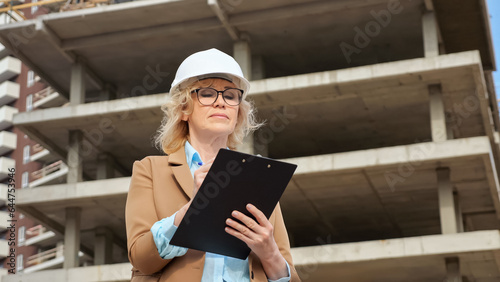 Senior woman entrepreneur makes notes on clipboard of building processes at construction site. Concept of working moments and production low angle shot