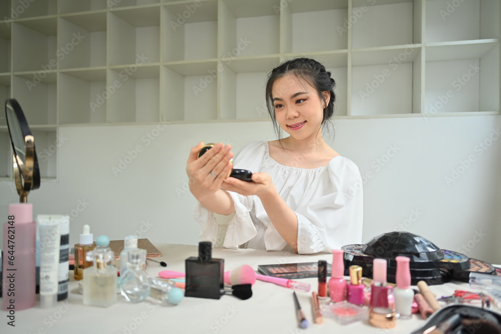 Smiling young woman holding compact powder and looking in mirror while doing daily makeup in dressing room