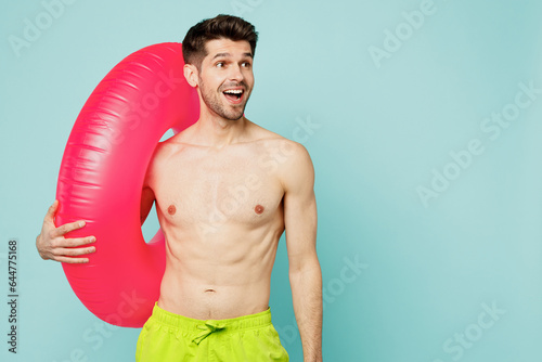 Young man wear green shorts swimsuit relax near hotel pool hold inflatable pink rubber ring look aside on area isolated on plain light blue cyan background. Summer vacation sea rest sun tan concept.