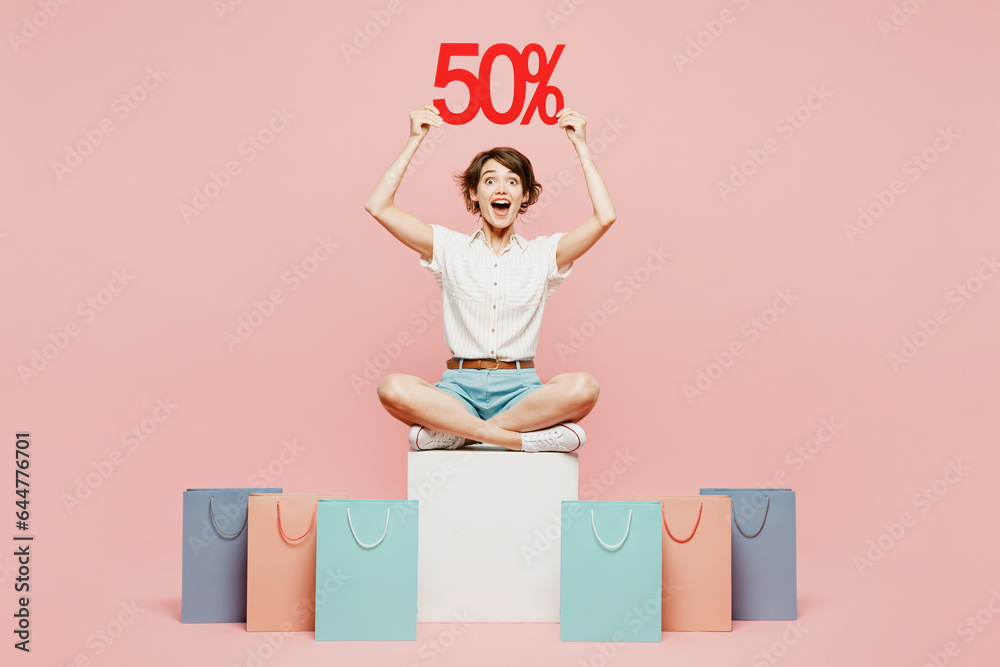 Full body surprised fun young woman wear casual clothes hold shopping paper package bags sitting hold 50 percent sale isolated on plain pink color background studio. Black Friday sale buy day concept. Full body surprised fun young woman wear casual clothes hold shopping paper package bags sitting hold 50 percent sale isolated on plain pink color background studio. Black Friday sale buy day concept.