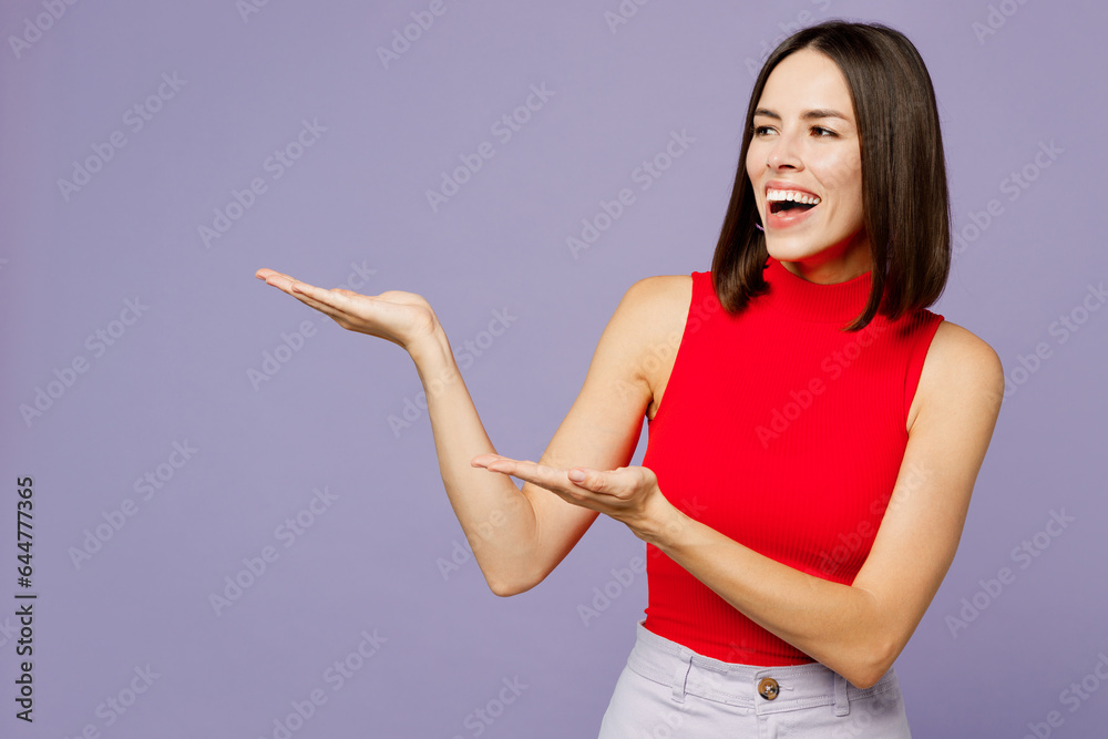 Young smiling woman she wearing red tank shirt casual clothes pointing hands arms aside indicate on workspace area copy space mock up isolated on plain pastel light purple background studio portrait.