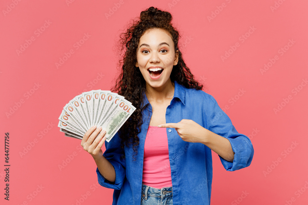 Φωτογραφία Young surprised woman of African American ethnicity wear blue shirt casual clothes hold in hand point finger on fan of cash money in dollar banknotes isolated on plain pastel pink background studio