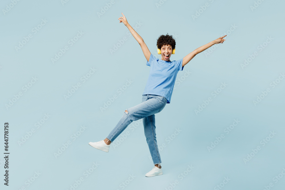 Full body young woman of African American ethnicity in t-shirt casual clothes listen to music in headphones raise up hands dance isolated on plain pastel light blue cyan background Lifestyle concept