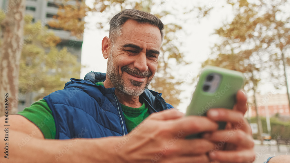 Fototapeta premium Close-up,happy mature man wearing casual clothes sitting on bench looking through information on smartphone