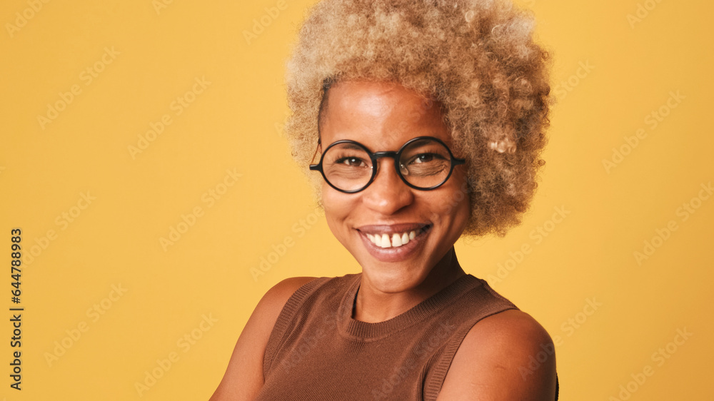 Close-up of girl in glasses, wearing brown top turning her head and looking at the camera with smile isolated on orange background
