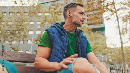 Mature man wearing casual clothes in wireless earphone enjoying music while sitting on park bench, bottom view, soft focus