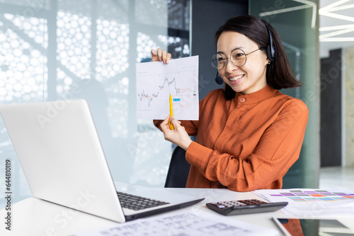 Online presentation remote meeting, asian woman with headset phone smiling and showing to laptop camera graph for good and successful achievement results, female worker inside office.