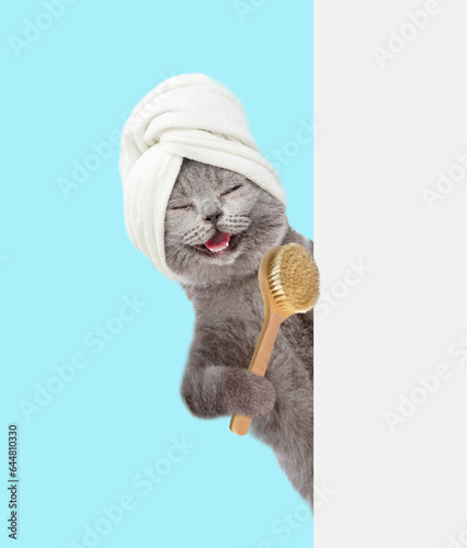 Fototapeta Naklejka Na Ścianę i Meble -  Happy cat with towel on it head looks from behind empty white banner and holds bath brush. isolated on blue background