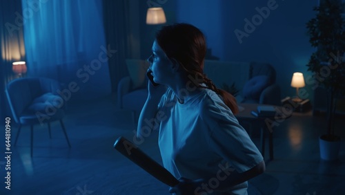 Shot of a young woman going around a dark room in search for burglars, intruders, thieves. She holds a baseball bat and a phone to call the emergency hotline.