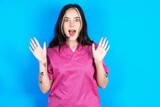 Optimistic Young caucasian doctor woman wearing pink medical uniform raises palms from joy, happy to receive awesome present from someone, shouts loudly,
