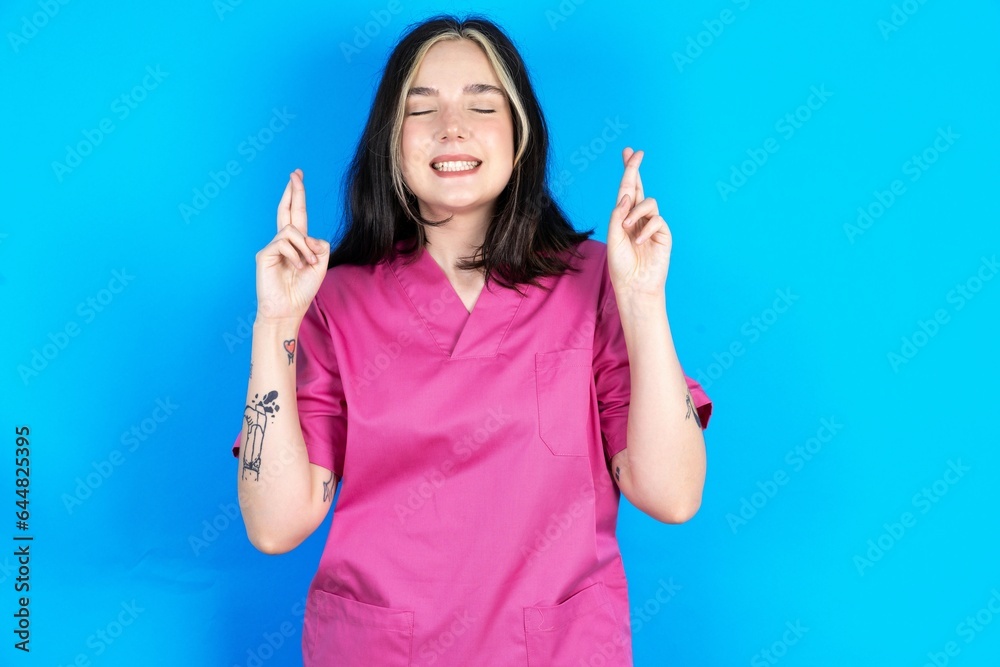 Young caucasian doctor woman wearing pink medical uniform has big hope, crosses fingers, believes in good fortune, smiles broadly. People and wish concept Young caucasian doctor woman wearing pink medical uniform has big hope, crosses fingers, believes in good fortune, smiles broadly. People and wish concept