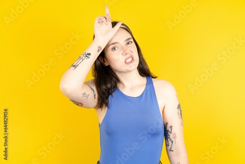 Beautiful woman wearing blue tank top making fun of people with fingers on forehead doing loser gesture mocking and insulting. photo