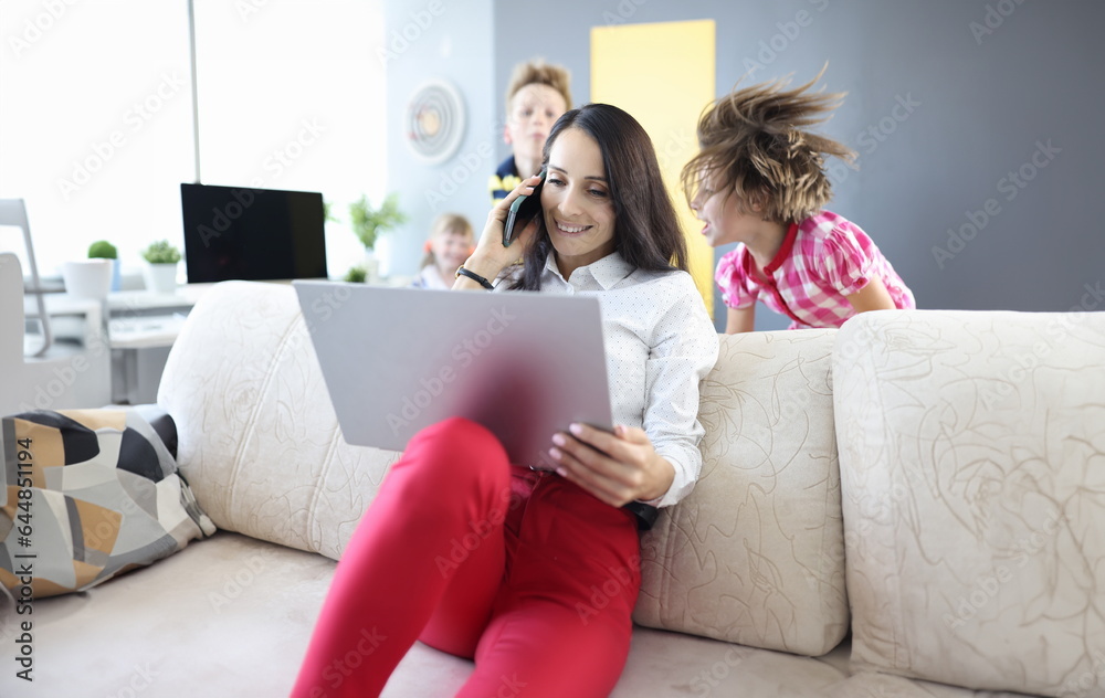 Woman is sitting on couch holding laptop and talking on smartphone next to the children are playing. Remote work in coronavirus in family concept Woman is sitting on couch holding laptop and talking on smartphone next to the children are playing. Remote work in coronavirus in family concept