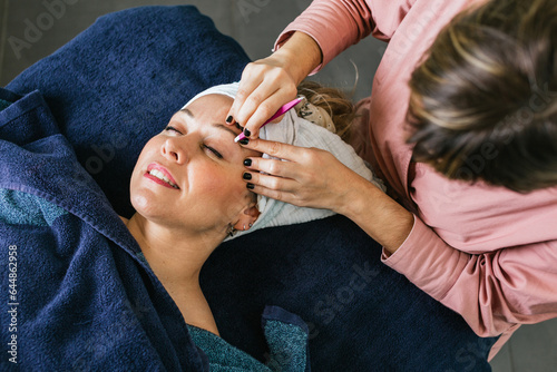 Unrecognizable beautician plucking eyebrows on face of woman in salon