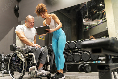 A determined individual in a wheelchair, working out in the gym with the guidance of a dedicated female fitness trainer who using a tablet to show a weekly workout plan. 