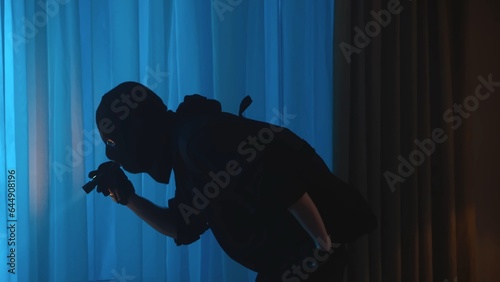 Shot of a burglar, intruder standing in the house, leaning forward with a flashlight on. The man is looking for valuables, jewelry. Night time, low, muffled light.