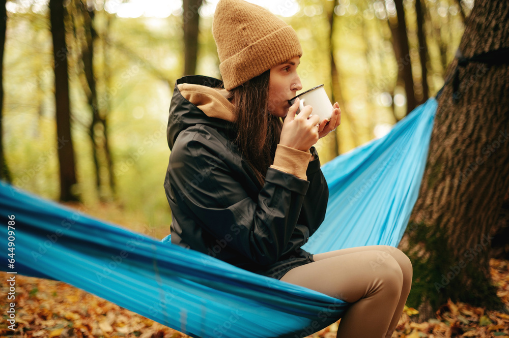 Woman sitting in hammock at autumn forest and drink tea or coffee Woman sitting in hammock at autumn forest and drink tea or coffee