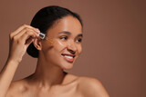 Smiling woman applying serum onto her face on brown background. Space for text