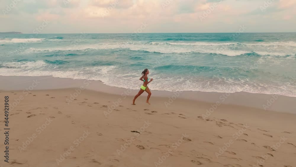 Drone shot. A young girl is jogging on the beach in the evening at sunset. Blue waves of the ocean wash the beach Drone shot. A young girl is jogging on the beach in the evening at sunset. Blue waves of the ocean wash the beach