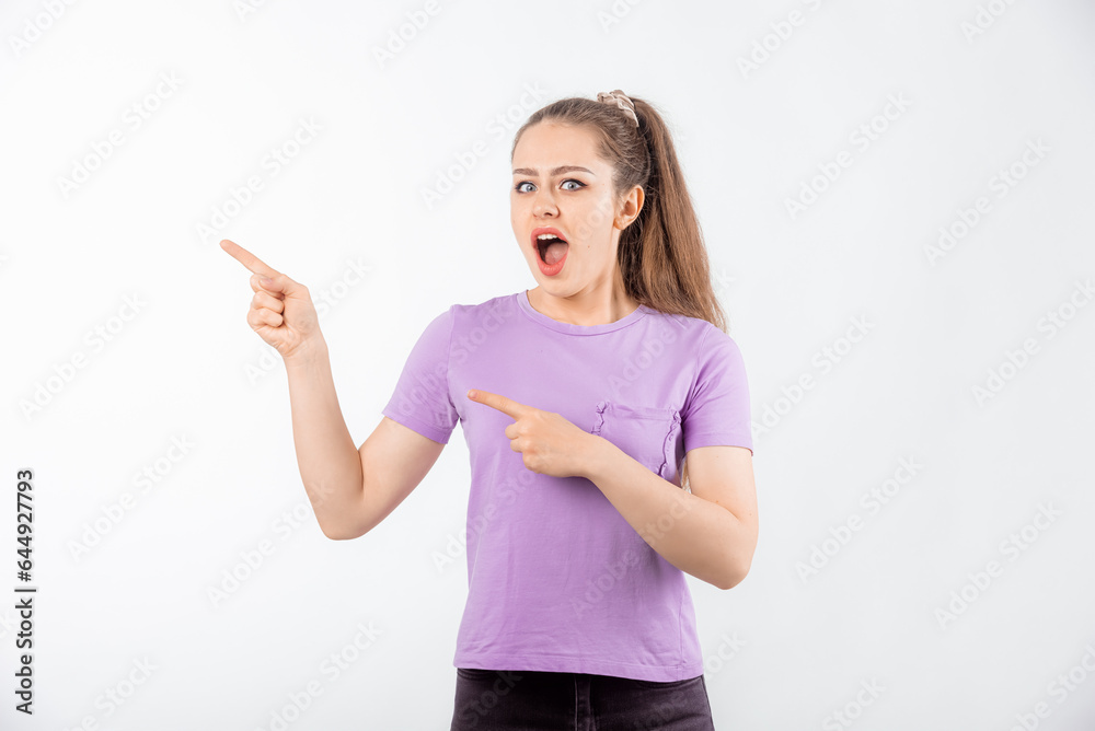 Surprised young woman with long hair points fingers at banner, shows great promo offer aside, looking amazed by promotion offer, stands in casual t shirt over white background Surprised young woman with long hair points fingers at banner, shows great promo offer aside, looking amazed by promotion offer, stands in casual t shirt over white background